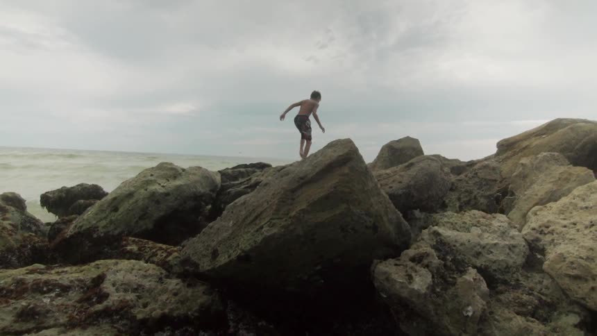 A boy in swim trunks climbs among rocks at the beach