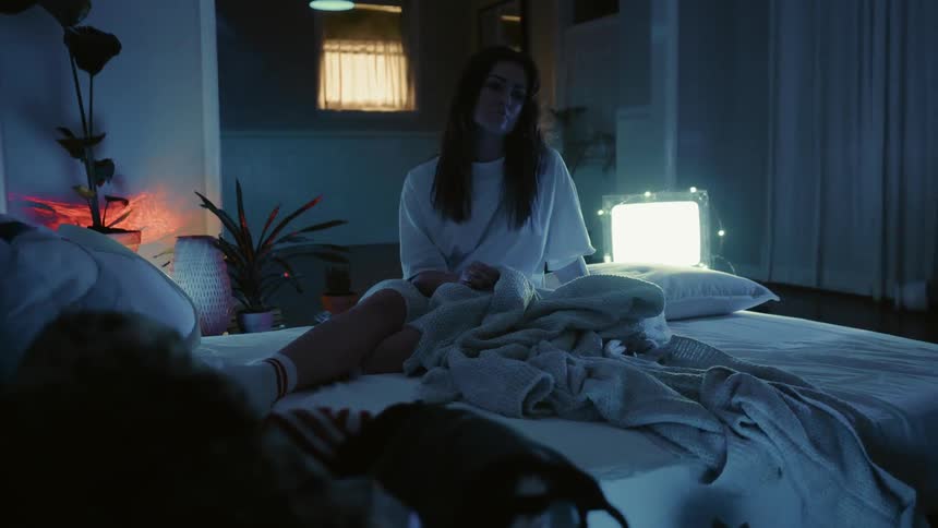 Woman sits on her bed as a red light shines around the room