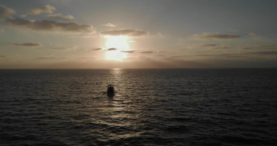 A small boat travels on the ocean at sunset with clouds in the sky