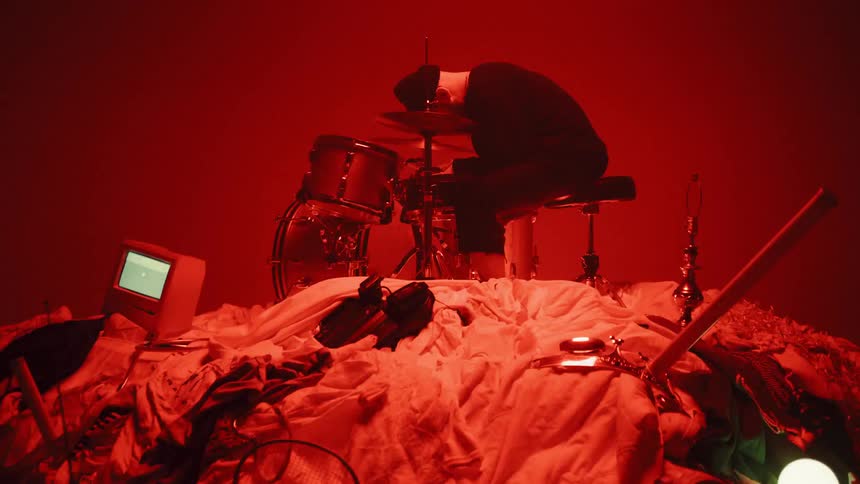 A  drummer sits on top of a pile of random objects