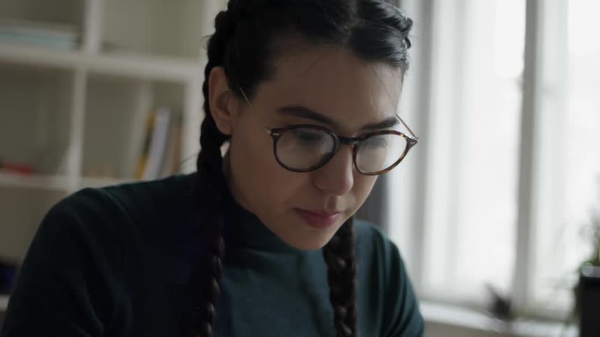 Woman wearing glasses and braids works near a window
