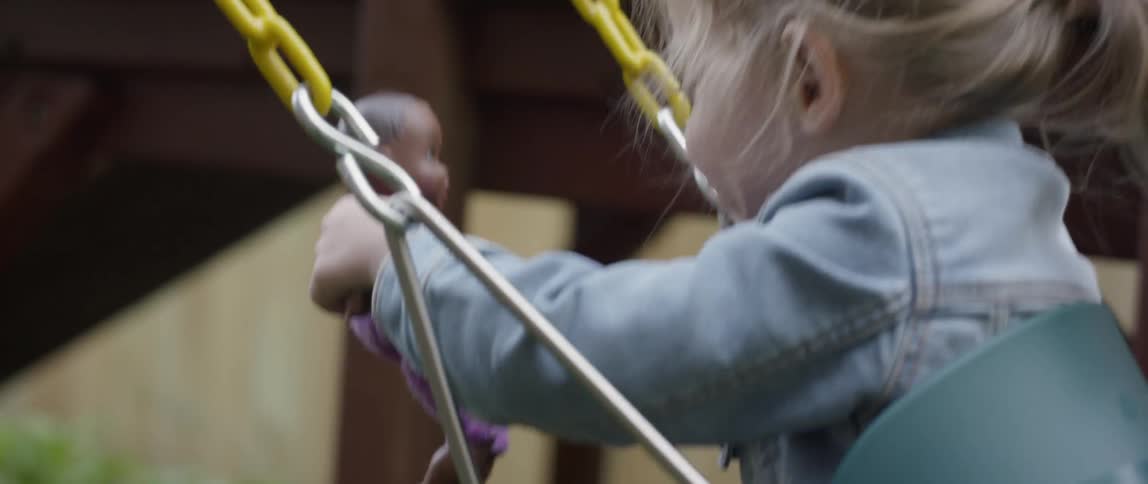 A little girl is holding her dolly as she swings on a swing set outside in a backyard in daylight