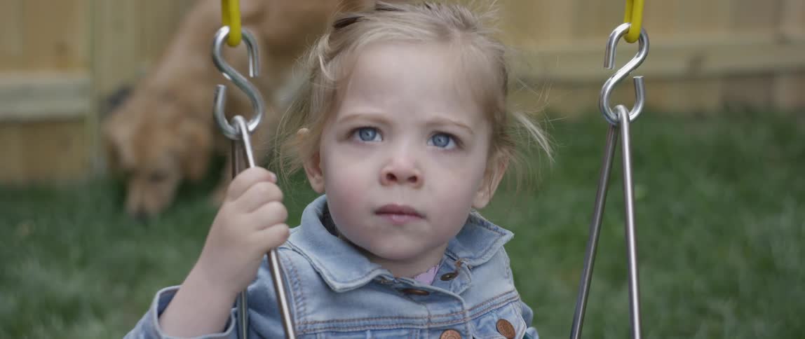 Little girl wearing a denim jacket swings on a swing as a dog walks behind her