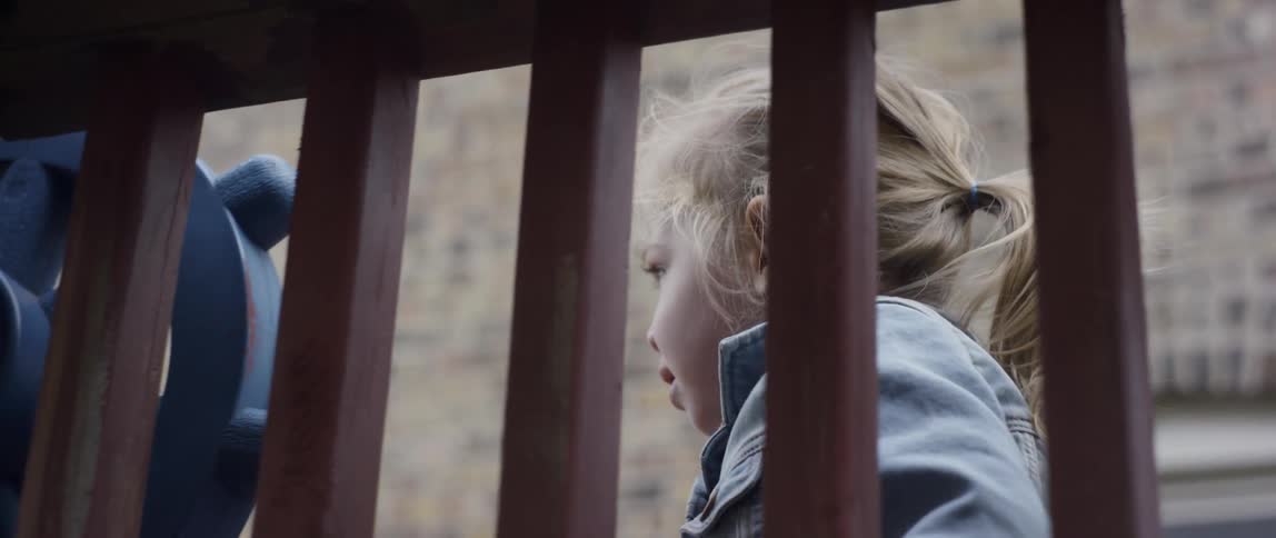 Little girl with blond hair walks near a brown railing holding her doll