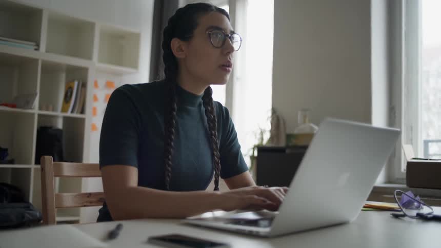 A woman types on a laptop and pauses to look up and think