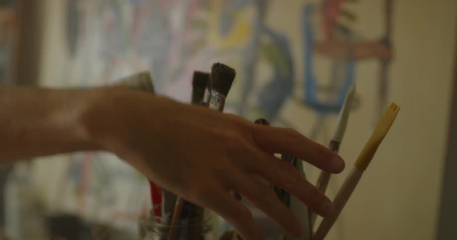 An artist is choosing palette knife from a container full of brushes inside in daylight