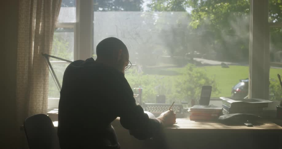 A man writes at a desk by the window