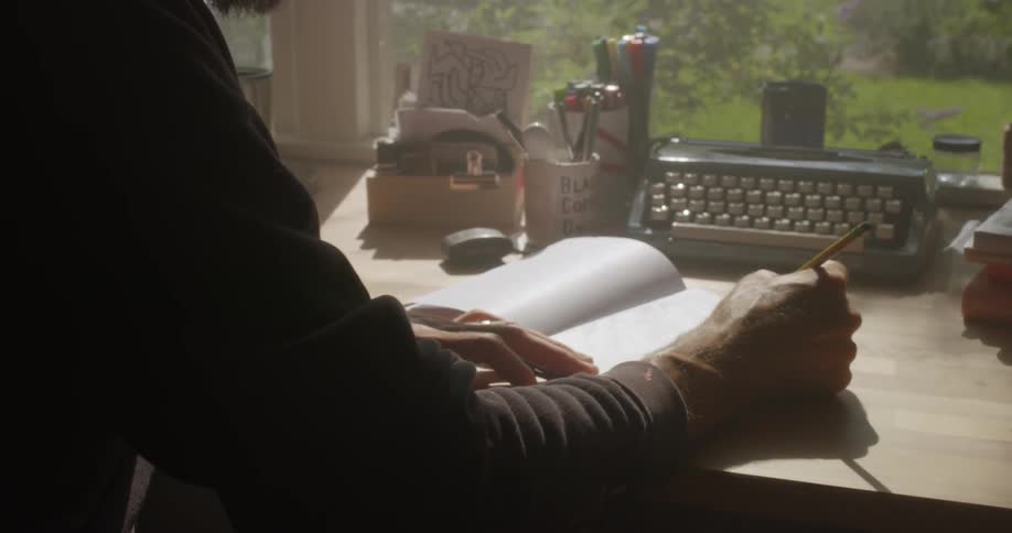 A man is sitting at a desk at home drawing in a notebook in daylight