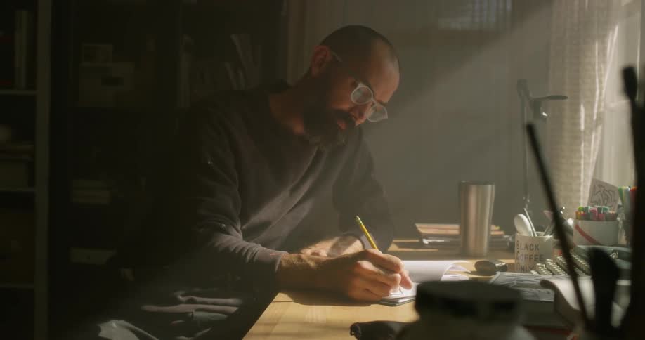 A man is sitting at a desk drawing in a notebook in daylight