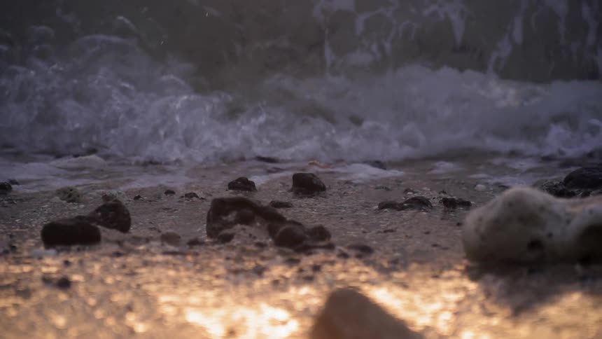A wave is splashing and rolling over rocks on a beach at sunset