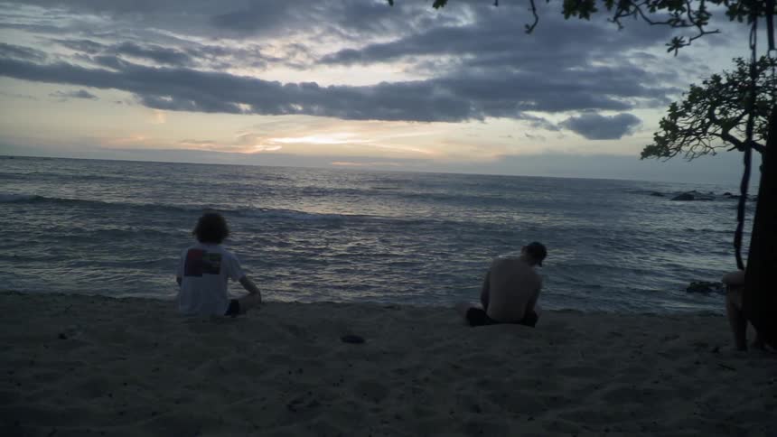 The two people sit on the sand at sunset