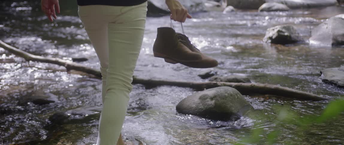 A woman wades in the river water while holding her shoes
