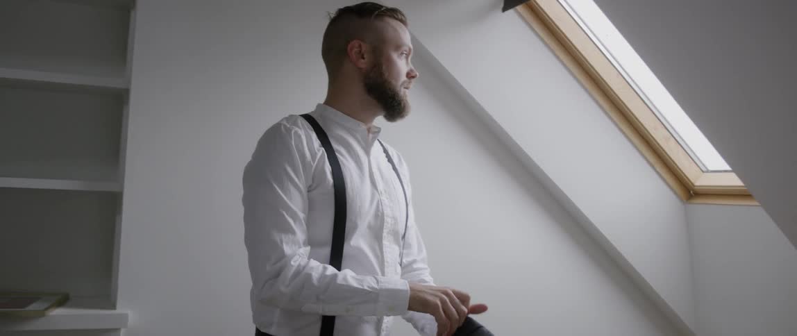 A man puts on a suit jacket next to an attic window