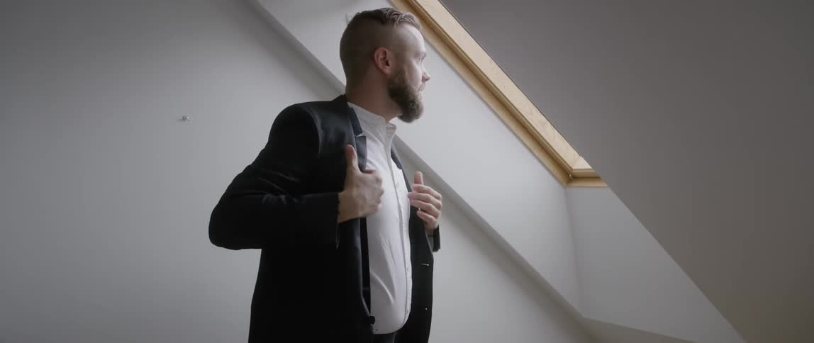 A man puts on a suit jacket next to an attic window