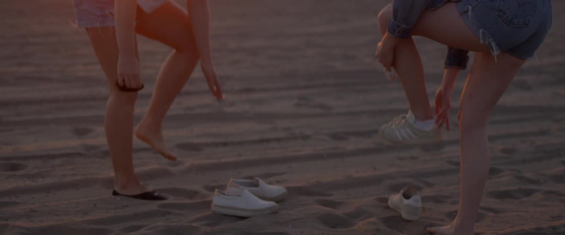 Two women take off their socks and shoes on a sandy beach at sunset
