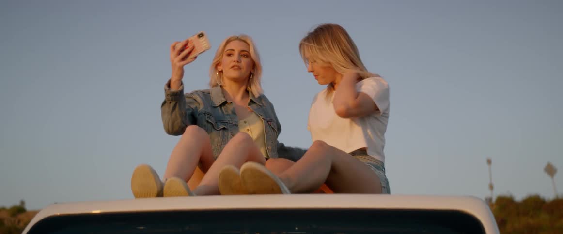 Two blonde women sit on the roof of a car and take a selfie