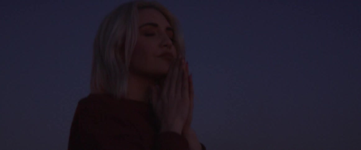 Blond haired woman clasps her hands as she stands under the night sky