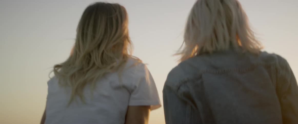 Two women climb up out of the sun roof and sit on the vehicle's roof at the beach