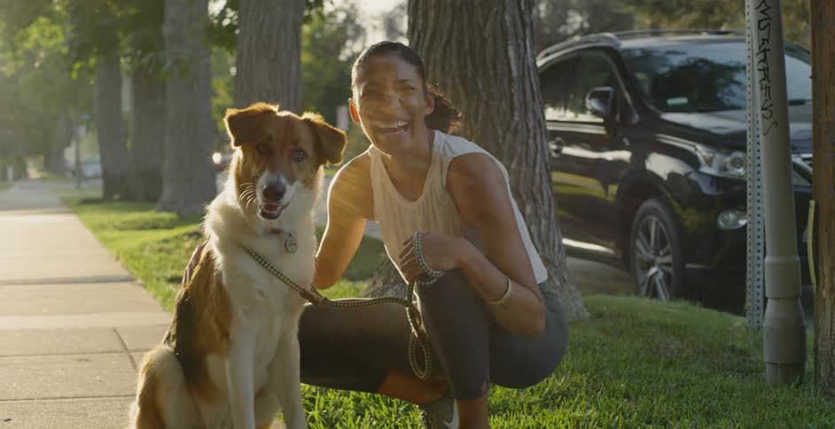 The woman is crouching in the grass petting her dog
