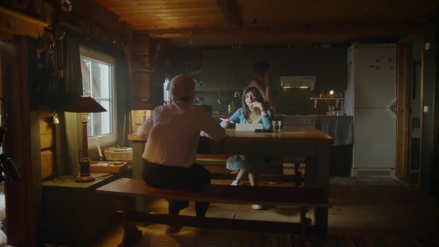 Two women sit at kitchen table talking as man paces in background