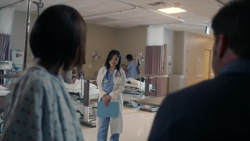 A woman is sitting with a man in a hospital and her doctor walks up