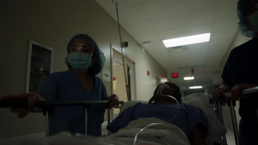 A woman with a oxygen mask lying on a bed as two people push her down a hall