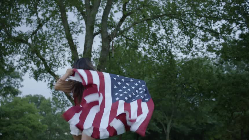 A girl runs through the grass toward trees while holding an american flag behind her