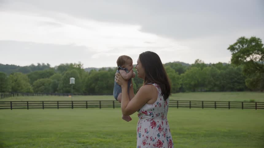A woman holds up and plays with a newborn in a rural field