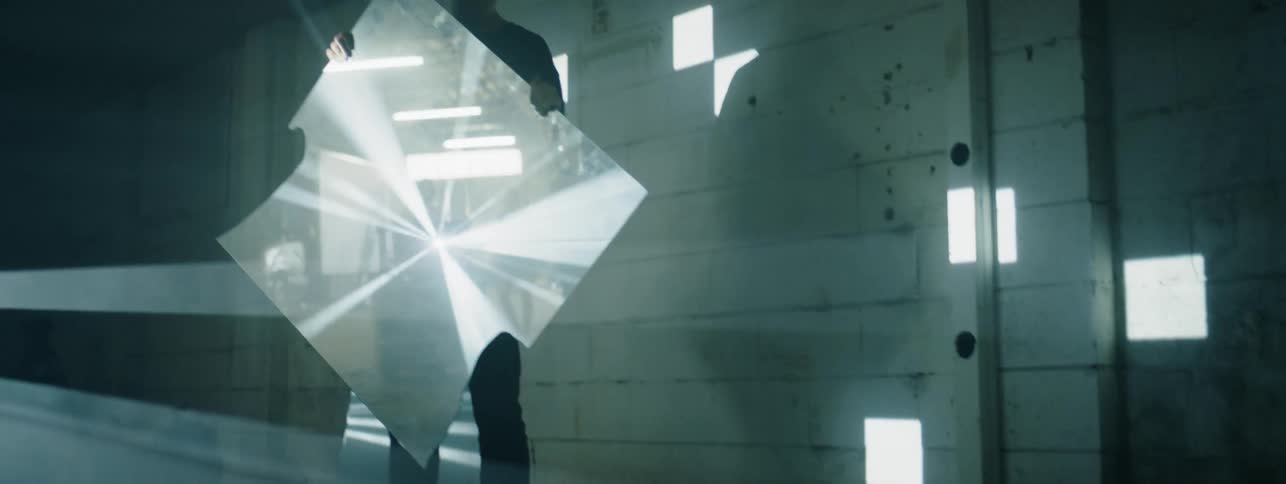 A man stands in a dark room holding a mirror as lights flash from across the room