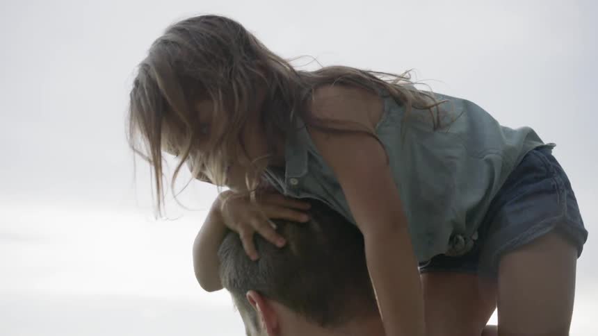 Young girl climbs up onto a man's shoulders under a cloudy sky