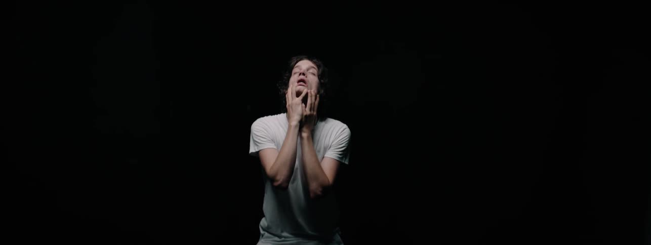 A boy sitting in a black room looks up while rubbing his face and neck
