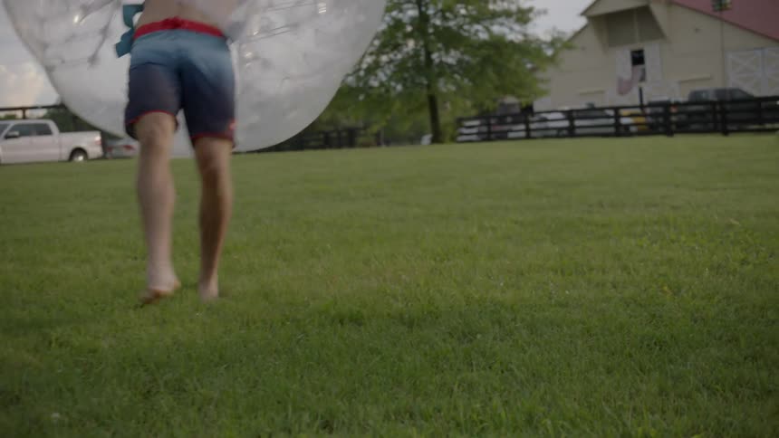 A man uses his phone to film two teenagers bouncing around in inflatable bubbles on the grass