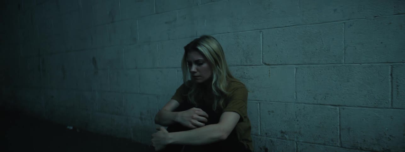 A woman is hugging her knees as she sits against a wall outside at night