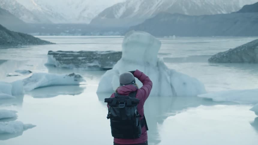 A woman wearing a backpack taking a photo of a glacier