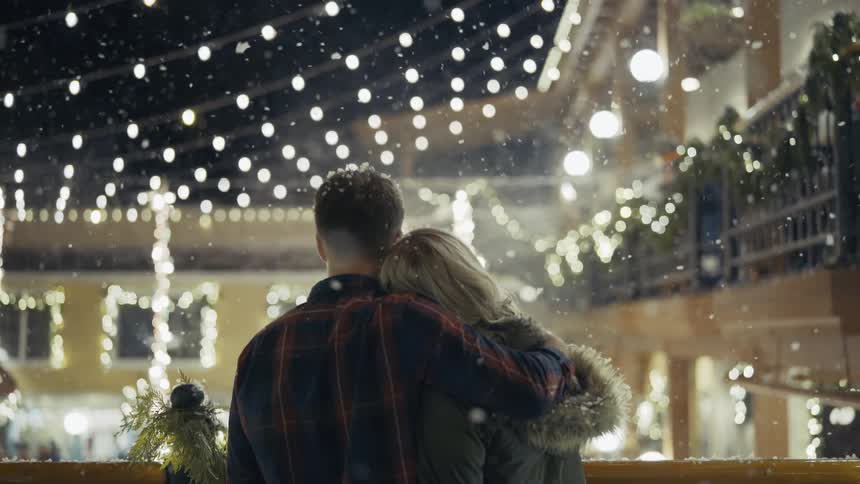 Man and woman cuddle as the snow falls and hanging lights shine on a building