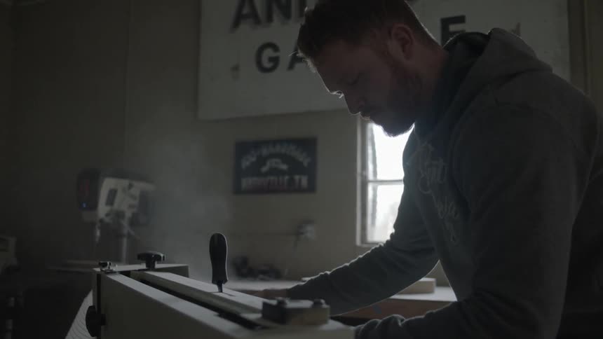A bearded man sanding a piece of wood with a table sander