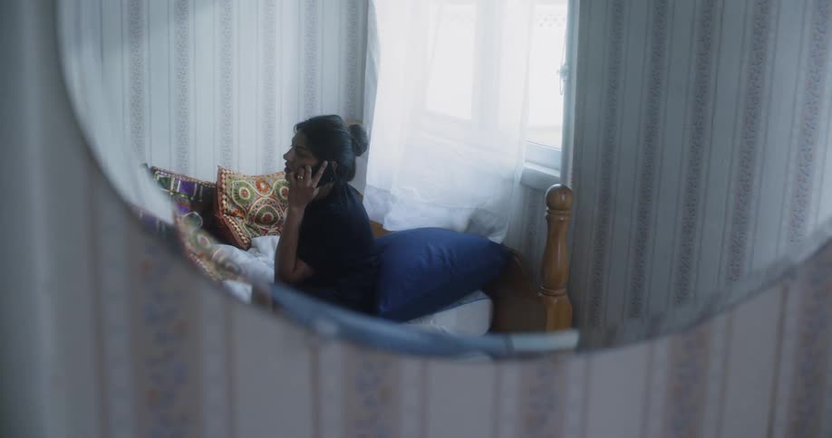 Woman sits on a bed and talks on the phone as a curtain flutters behind her