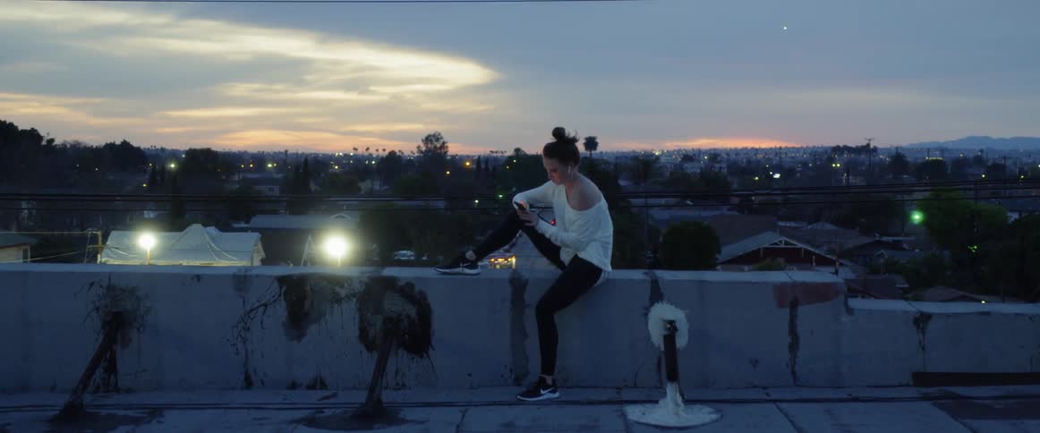 A female sitting on a ledge of a rooftop building at night fall on her phone