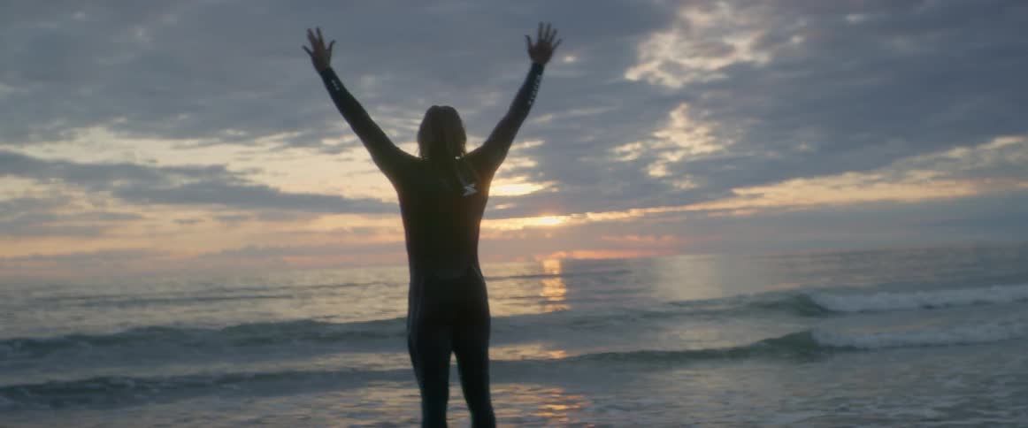 A man raises his arms and touches his hands together then lowers them as he stands by the ocean with a surfboard at his feet