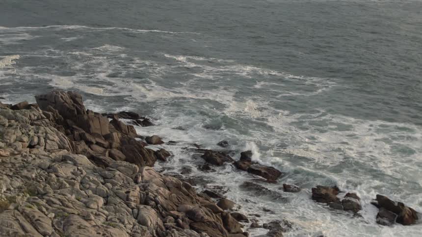 A rocky cliffside has white topped waves crash around  it's rocky base