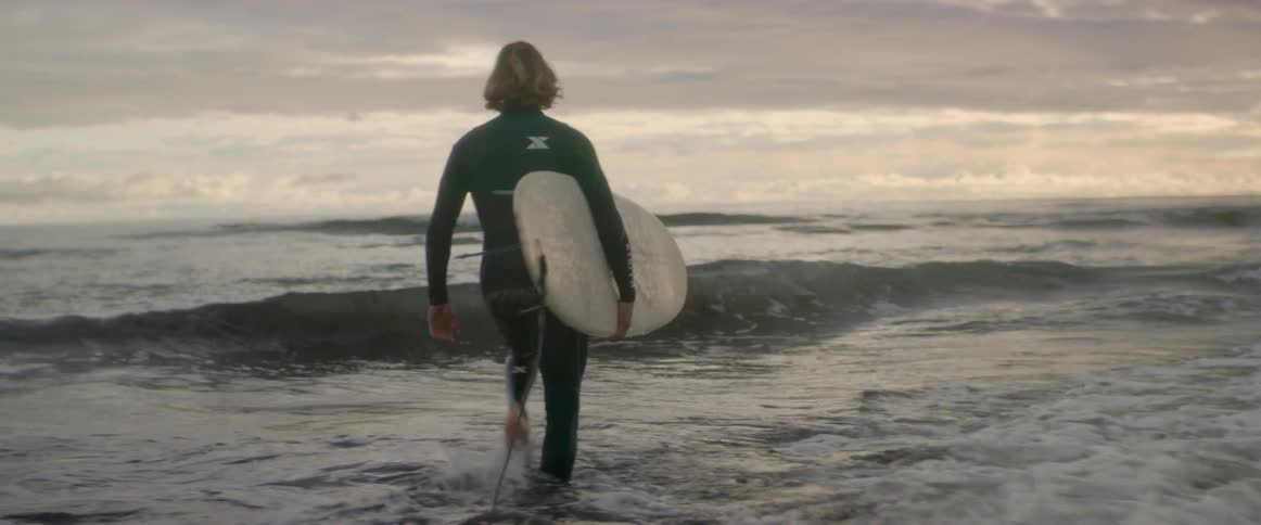 Man walking on a beach through the waves while holding a surfboard