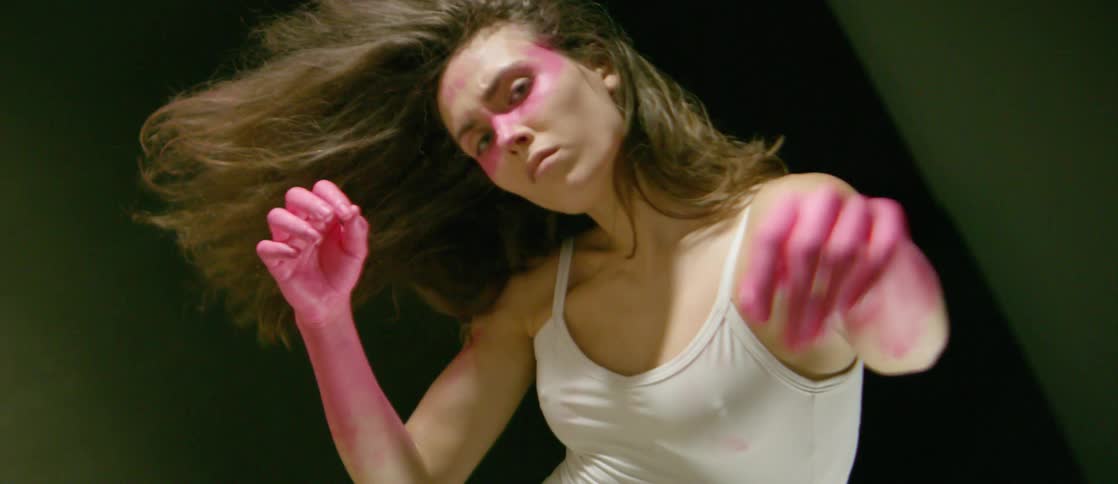 A woman does some unique movements with pink paint on her face and arms