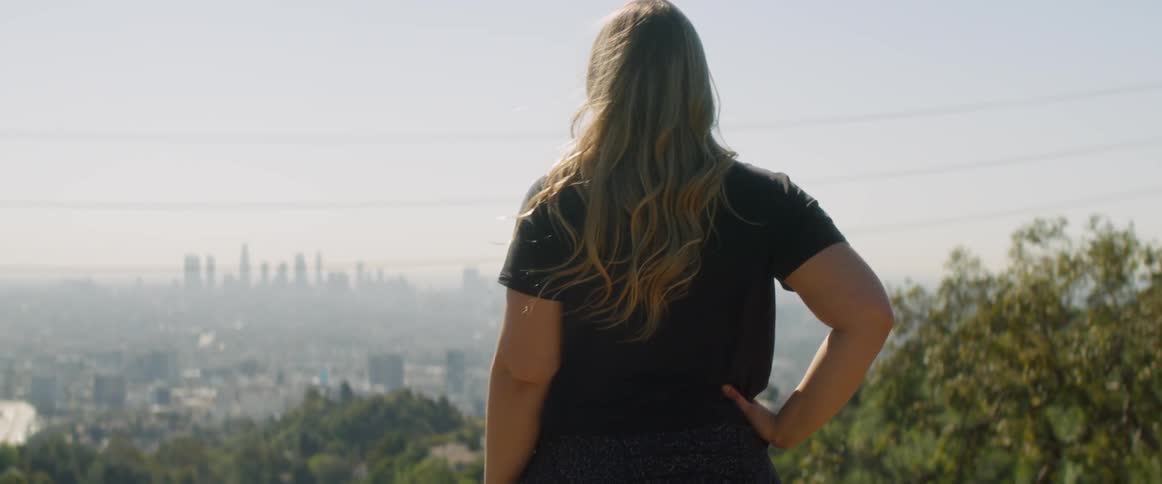 A woman stands on a large hill looking down at the city below in the sunrise