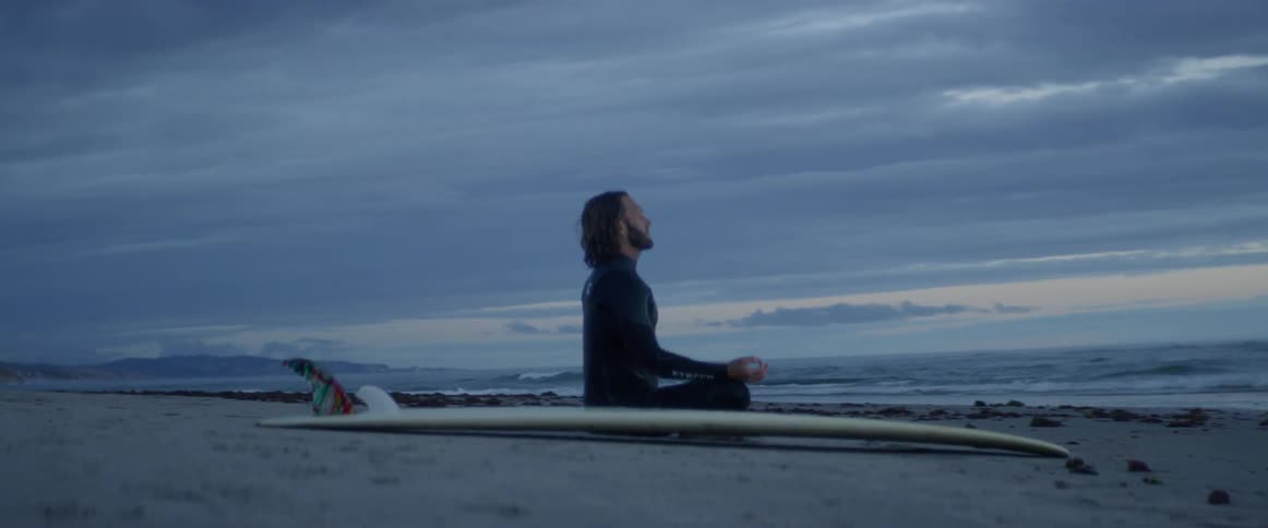 Man sitting on a beach at sunrise next to his surfboard