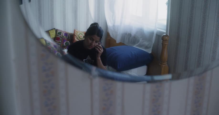 A woman sits on a bed talking on the phone as a curtain flutters in the wind behind her