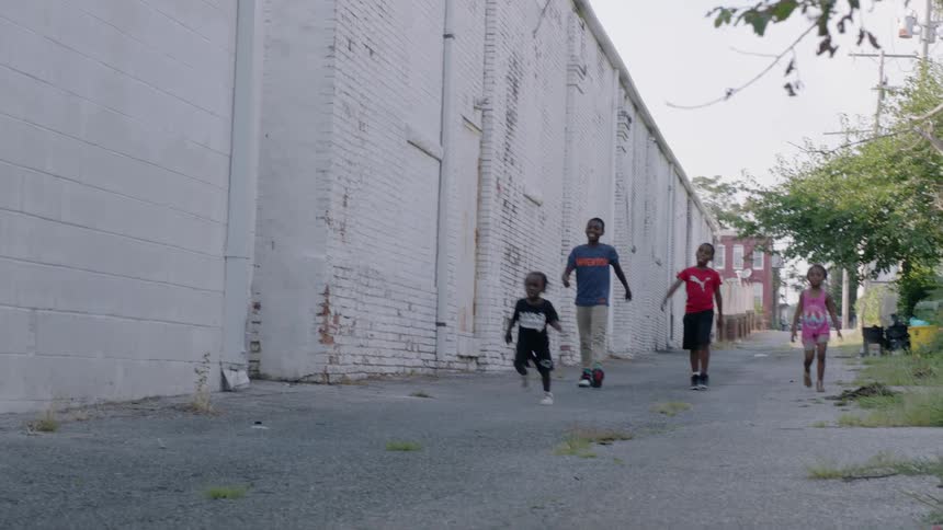 A couple of children young and older are walking down an alley with a large white wall by them