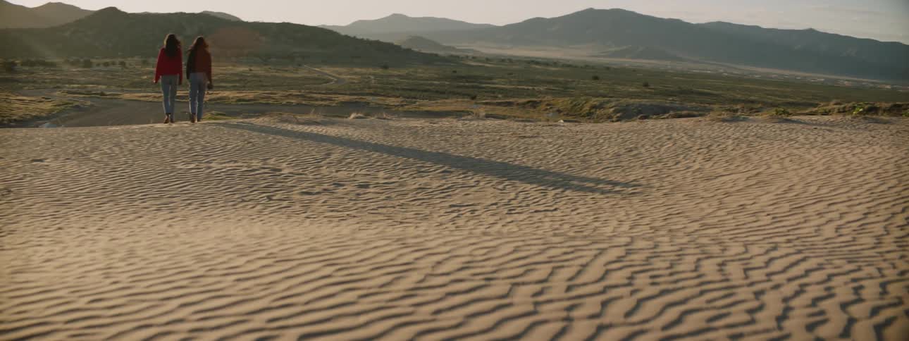 Two women walk through a desert at sunrise