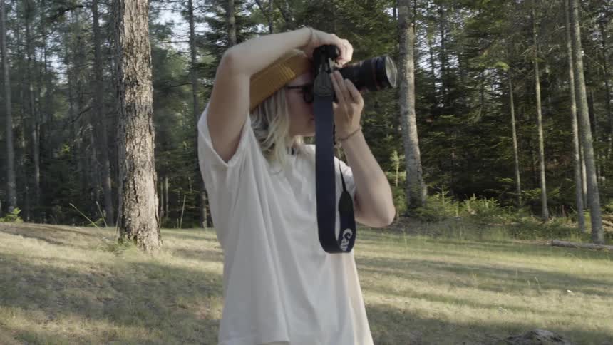 A young man with long hair looks through a camera while standing in a forest