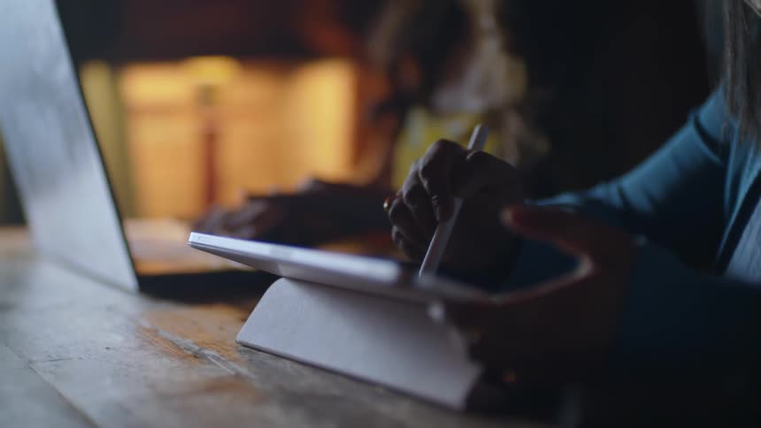 A woman writes on a tablet with a stylus while another woman types on a keyboard behind her