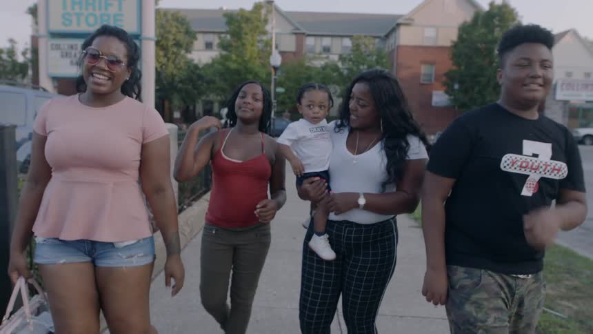 Four women smile and walks on the sidewalk in a city and two women hold a toddler